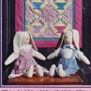 Puede incluir: Un folleto rosa con el título "Bunnies and Baskets" presenta dos muñecos de conejos folk teñidos con té y un edredón a juego. El edredón tiene diseños de cestas en rosa, azul y amarillo. El folleto incluye el texto "Erica's Quilts".