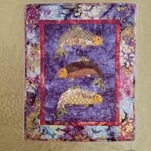 May include: A fabric art piece titled "Fish Tales" featuring three stylized fish in shades of brown, gold, and tan against a purple background. The artwork measures 26.5 inches by 33 inches and is framed with a red border.