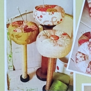 May include: A collage of images showcasing handmade pincushions. The pincushions are round, with fabric patterns in shades of red, yellow, and white. Some are on stands, others in mugs, and some are in a dish. The text "Fig Tree threads" and "Petite Gateau" are visible.