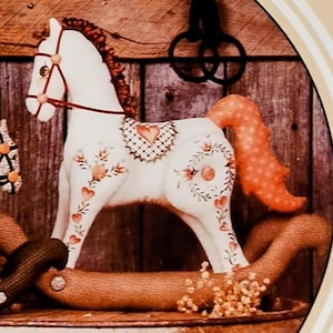 May include: A craft pattern titled "Country Charm" featuring two jointed hobby horses. The horses are white and tan with orange manes and tails, set against a rustic wooden backdrop. The pattern includes directions for 8" and 12" fabric horses.