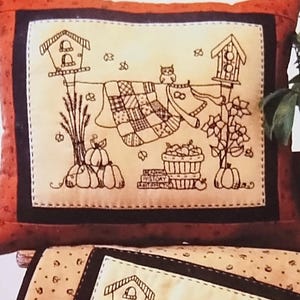 May include: A sewing pattern cover titled Blueberry Backroads featuring an orange pillow and a tan quilted mat, both displaying a line-art illustration of birdhouses, a quilt on a clothesline, pumpkins, and an owl. A sign reads Autumn in Grandma's BackYard.
