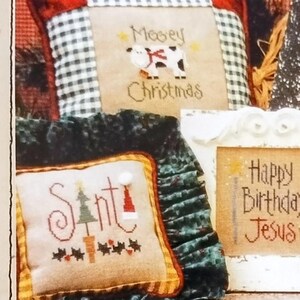 May include: A collection of cross-stitch patterns featuring holiday-themed designs. The image includes framed samplers with phrases like "Merry Christmas," "Just add snow," and "Happy Birthday Jesus." The title "Tiny Tidings VI" is also visible.