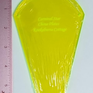 May include: A transparent yellow plastic quilting template, approximately 5 inches long, wrapped in clear plastic. The template has a unique shape and features the text "Carnival Star China Plates Kookaburra Cottage". A ruler is placed next to the template.