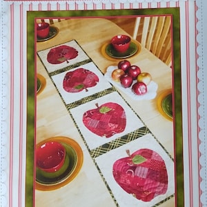 May include: A patchwork apple table runner with four large red apple designs. The runner is on a wooden table, with red bowls and apples on the table. The image has the text "Shabby Fabrics" and "Patchwork Apple Table Runner".