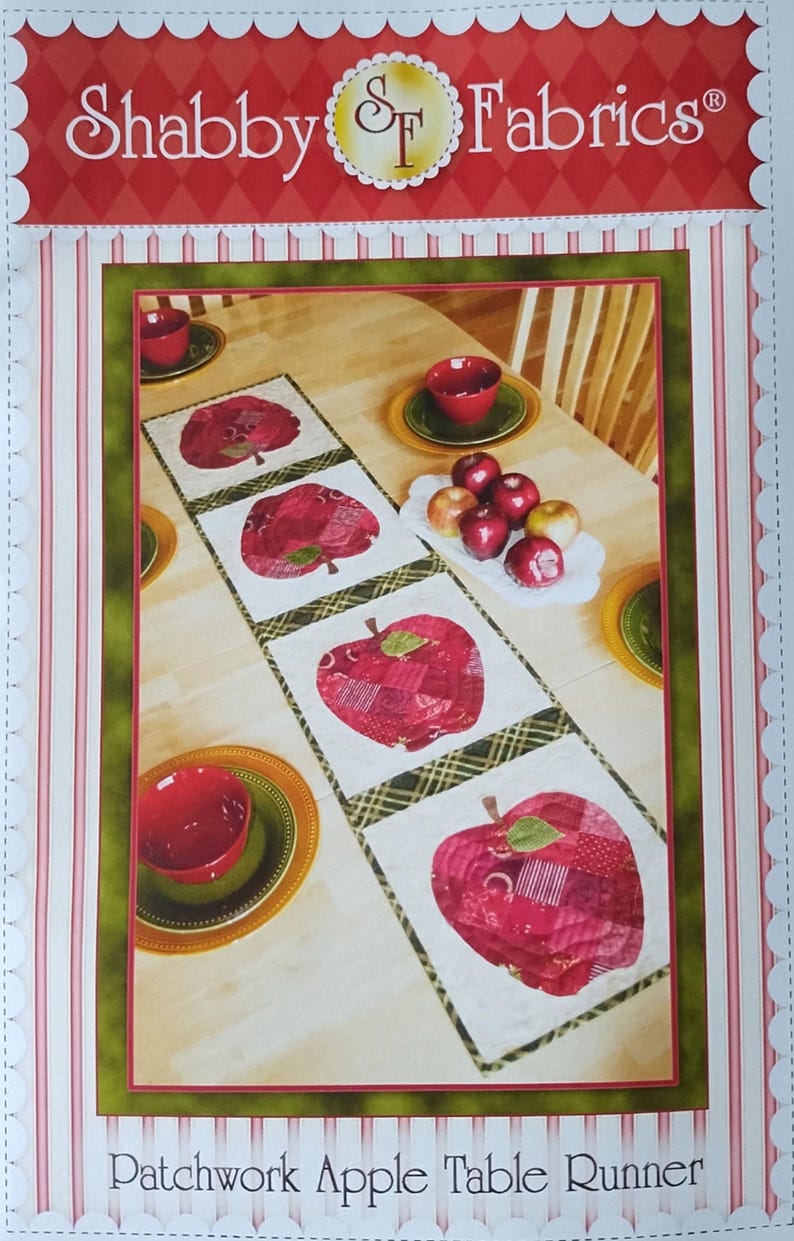 Op de afbeelding: Een patchwork tafelloper met rode appelontwerpen. De loper ligt op een houten tafel, met rode kommen en appels. De afbeelding bevat de tekst "Shabby Fabrics" en "Patchwork Apple Table Runner".