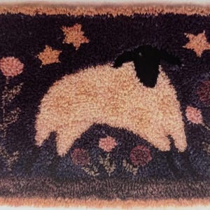 May include: A rectangular rug with a sheep design. The sheep is beige with a black head and is surrounded by stars and flowers. The background is dark blue, and the border is a light brown.