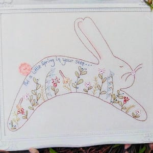May include: Embroidered artwork in a white frame. The artwork features a rabbit silhouette filled with floral and plant designs. Text reads, "Put a little spring in your step..." in a cursive font.