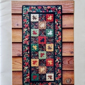 May include: A paper foundation pattern for a quilted table runner. The design features colorful leaf squares and a floral border. The text reads "Tumbling Leaves Runner" and "MH Designs". The background is a wood grain.