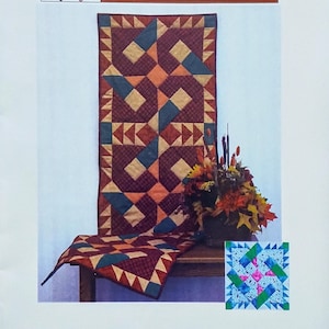May include: A brochure for "Table Relish" by TyQuilts Designs. The image features a quilted table runner with geometric patterns in shades of red, orange, blue, and tan. A small floral arrangement and a sample quilt block are also visible.