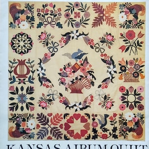 May include: A cream-colored quilt with a floral design. The quilt features various floral patterns, including roses, bows, and birds, in shades of pink, red, and blue. The text "Osage County Quilt Factory" and "Kansas Album Quilt" are at the top and bottom.