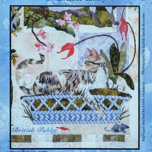 May include: A quilt block titled "My Cats Garden" featuring a tabby cat in a woven basket with floral accents. The quilt block is labeled "#8 British Tabby" and includes the text "Maggie Walker Design".