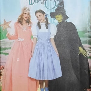 May include: Simplicity sewing pattern featuring costumes from The Wizard of Oz. The image shows three characters: Glinda in a pink gown, Dorothy in a blue dress, and the Wicked Witch of the West in a black cloak and hat.