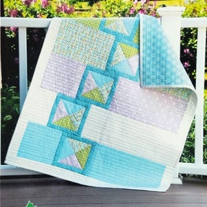 May include: A colorful quilt with a "Jack's Boxes" pattern, featuring squares and triangles in shades of blue, green, and lavender. The quilt is displayed outdoors on a white railing, with a green and floral background. The quilt is made by Robinson Pattern Company.