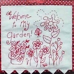 May include: A light blue fabric square with a red polka dot border. The center features a hand-drawn garden scene with flowers, leaves, and the text "Welcome to My Garden!" in red thread.