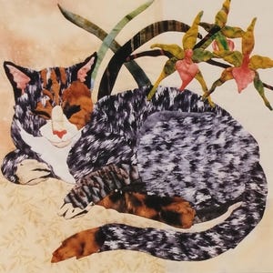 May include: A fabric art piece depicting a sleeping calico cat with a white chest and nose, and orange and black markings. The cat is curled up next to a sprig of yellow and pink orchids. The words "American Tabby" are written in cursive.
