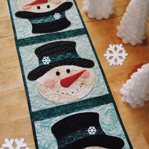 May include: A rectangular quilted table runner featuring four repeating snowman head appliques. Each snowman wears a black top hat with a small white snowflake accent, a red carrot nose, and pink cheeks, set against a light green patterned background.