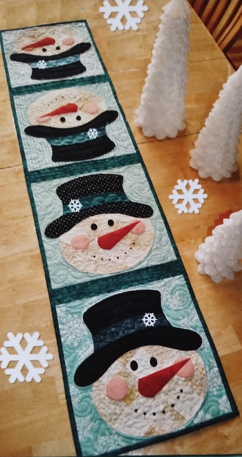 May include: A rectangular quilted table runner featuring four repeating snowman head appliques. Each snowman wears a black top hat with a small white snowflake accent, a red carrot nose, and pink cheeks, set against a light green patterned background.