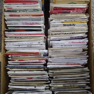 May include: A cardboard box filled with a collection of vintage sewing patterns. The patterns are from brands like Simplicity, Butterick, McCall's, and Vogue. The patterns have various colors and designs, and are stacked neatly.