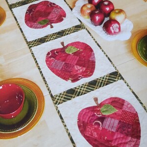 Op de afbeelding: Een gewatteerde tafelloper met grote rode appelontwerpen. De appels zijn gemaakt van verschillende rode stoffen en hebben groene bladeren. De loper is afgezet met een ruitpatroon. Rode kommen en borden met appels zijn ook zichtbaar.