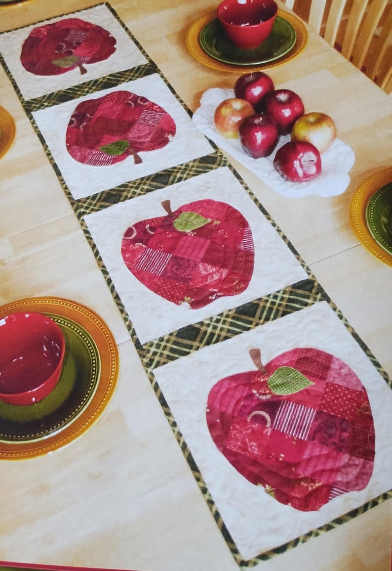 Op de afbeelding: Een gewatteerde tafelloper met grote rode appelontwerpen. De appels zijn gemaakt van verschillende rode stoffen en hebben groene bladeren. De loper is afgezet met een ruitpatroon. Rode kommen en borden met appels zijn ook zichtbaar.