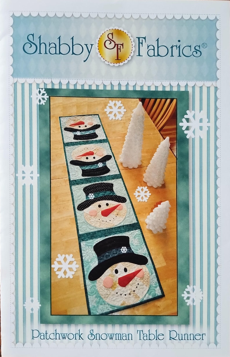 May include: A rectangular patchwork table runner featuring four snowman faces wearing black top hats. The snowmen have orange carrot noses, black eyes, and pink cheeks. The runner is set against a light blue background with white snowflake cutouts.