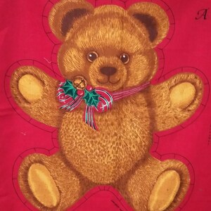 May include: A fabric panel featuring a printed brown teddy bear on a solid red background. The bear has a textured fur appearance, dark eyes, and a nose. A festive ribbon with green holly leaves, red berries, and a small gold bell is at its neck.
