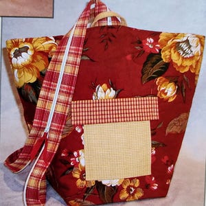May include: A red tote bag with a floral pattern in yellow and white. The bag features a plaid zipper and a beige square patch. The handles are made of a light brown material.