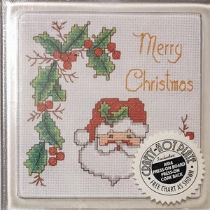 May include: A cross-stitch Christmas design featuring Santa Claus and holly with red berries. The words "Merry Christmas" are stitched in gold thread. Includes a "Crafty-Hot Plate" with a press-on board and cork back.