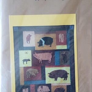 May include: A craft kit featuring a patchwork design with various pig illustrations. The word "Pigs" is at the top, with "machine appliqué designs" below it. The design includes pigs in different colors and poses, set against a colorful background.
