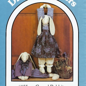 May include: Image of a pattern for Grand Rabbits, featuring two plush rabbit figures. One rabbit is hanging, wearing a floral dress and pants. The other rabbit is seated, wearing a purple outfit. The text reads "Dream Spinners" and "#155 - Grand Rabbits".