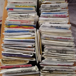 May include: A collection of vintage sewing patterns from brands like Simplicity, McCall's, and Butterick. The patterns are stacked in neat rows, with visible text including pattern numbers and brand names. The color palette is primarily white, with accents of red, blue, and yellow.