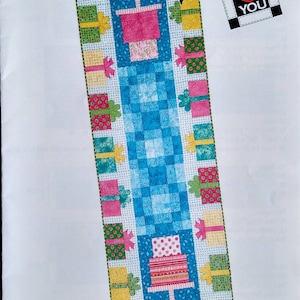 May include: A colorful Party Time table runner pattern. The design features a central blue checkered pattern with a pink cake and gift boxes. The table runner measures 26" x 74". Includes the text "from ME to YOU".