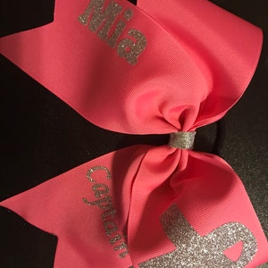May include: Pink cheer bow with silver glitter lettering that says "Mia" and "Captain". A silver glitter ribbon is in the center of the bow.