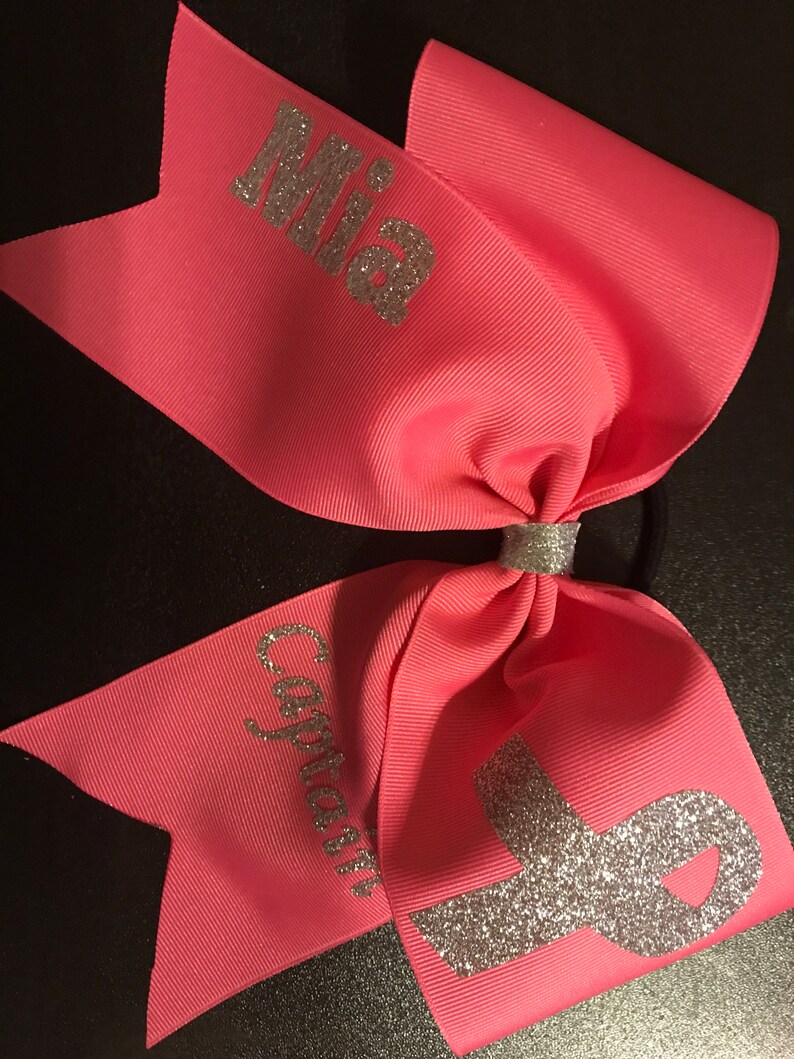 May include: Pink cheer bow with silver glitter lettering that says "Mia" and "Captain". A silver glitter ribbon is in the center of the bow.