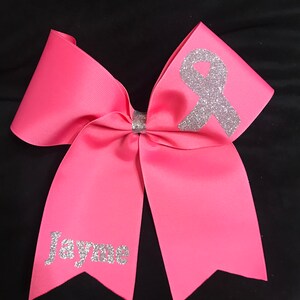 May include: Pink cheer bow with a silver glitter ribbon and the name "Jayme" in silver glitter.