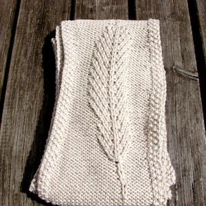 Könnte beinhalten: Ein gefalteter, cremefarbener Strickschal mit einem zentralen blattartigen Design. Der Schal ist aus einem weichen, strukturierten Garn gefertigt und scheint handgestrickt zu sein. Der Schal ist ordentlich auf einer Holzoberfläche gefaltet.