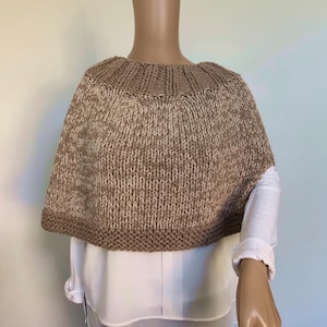 Op de afbeelding: Een bruin en wit gebreid poncho met een brede, geribbelde halslijn. De poncho is gemaakt van een zacht, getextureerd garen en heeft een ontspannen pasvorm.