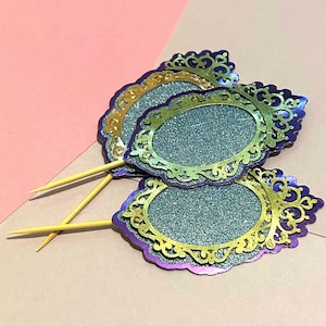 May include: Three decorative cupcake toppers with an oval glitter center and ornate gold and purple filigree edges. Each topper has a wooden pick attached for easy insertion into baked goods. The toppers are arranged on a pink and beige background.
