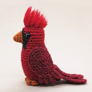 May include: A crocheted cardinal bird toy with a red body, black eyes, and an orange beak. The bird has a fluffy red crest on its head.