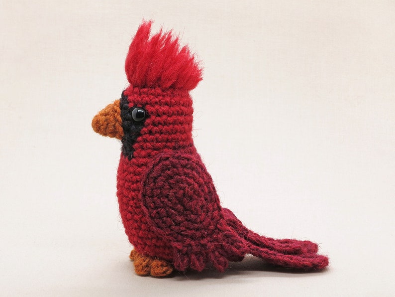 May include: A crocheted cardinal bird toy with a red body, black eyes, and an orange beak. The bird has a fluffy red crest on its head.