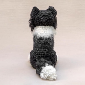 May include: A black and white crocheted dog toy with a fluffy tail. The dog is sitting with its back to the camera.
