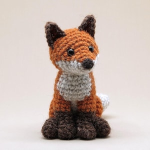 Pode incluir: Um brinquedo de pelúcia de raposa de crochê com pelo laranja, branco e marrom. A raposa está sentada com a cabeça inclinada para cima e a cauda enroscada atrás dela.