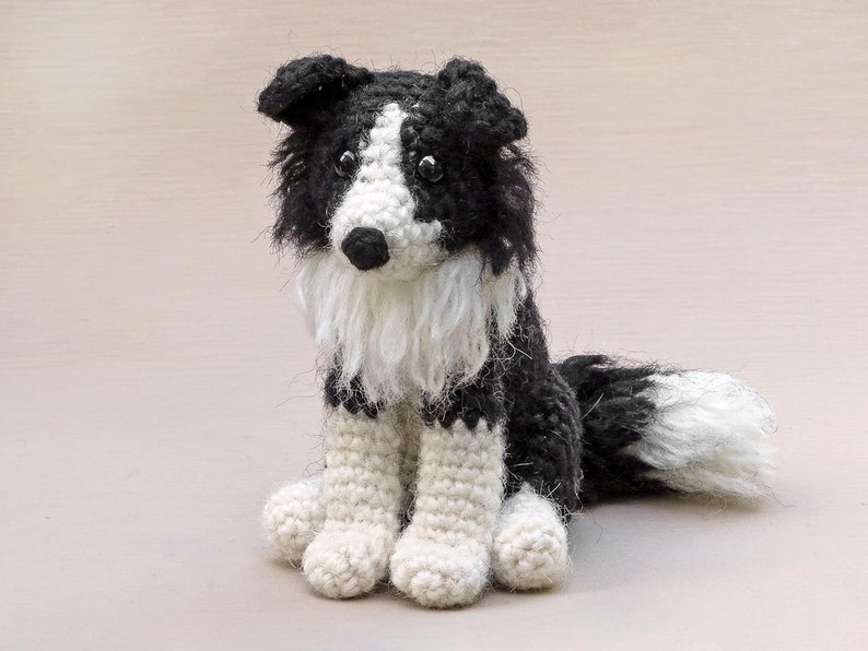 May include: A crocheted black and white border collie dog sitting with its tail curled to the side. The dog has a white patch on its chest and white fur around its face.