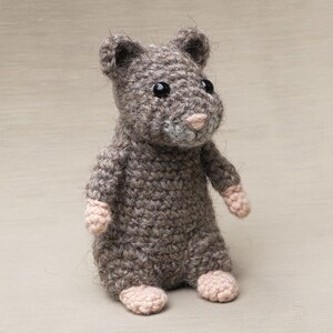 May include: A small, gray crocheted hamster toy with pink ears and nose. The toy has black eyes and is sitting on a light brown surface.