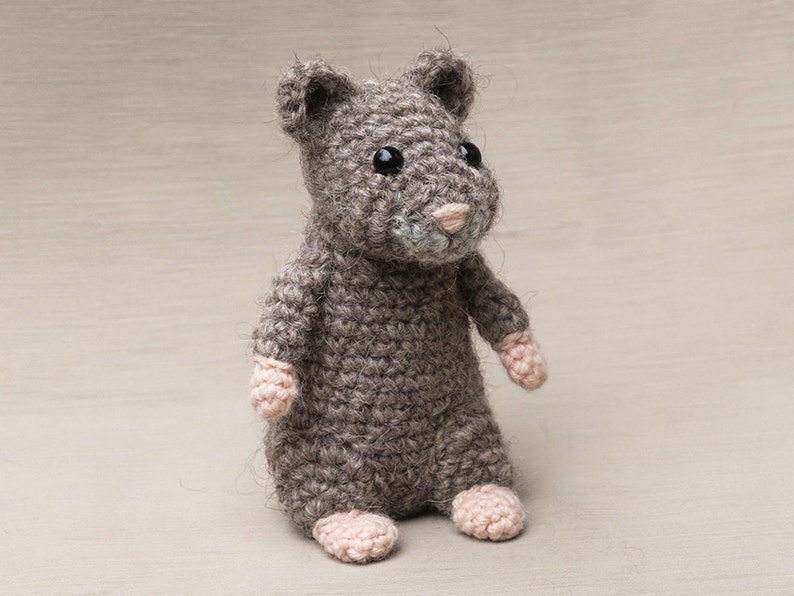 May include: A small, gray crocheted hamster toy with pink ears and nose. The toy has black eyes and is sitting on a light brown surface.