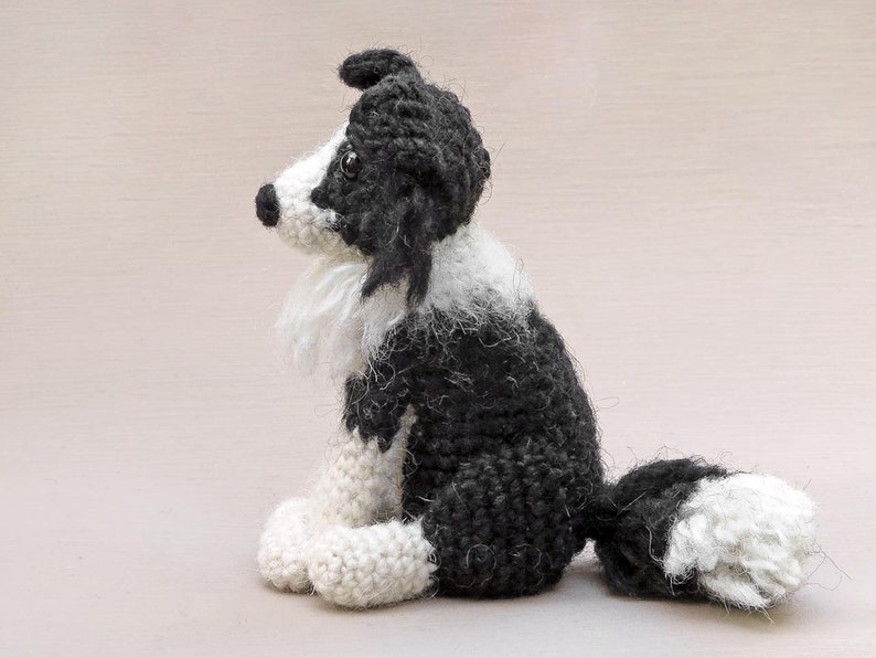 May include: A crocheted black and white border collie dog sitting with its tail curled behind it. The dog is made of yarn and has a fluffy white chest and tail.