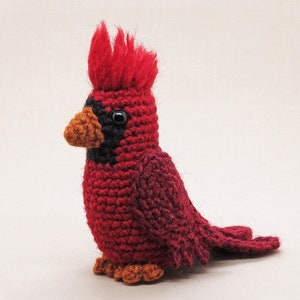 May include: A crocheted cardinal bird toy with a red body, black eyes, and an orange beak. The bird has a red and black crest on its head.