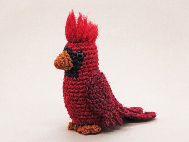 May include: A crocheted cardinal bird toy with a red body, black eyes, and an orange beak. The bird has a red and black crest on its head.