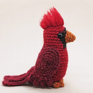 May include: A crocheted cardinal bird toy with a red body, black eyes, and an orange beak. The bird is sitting on a white surface.