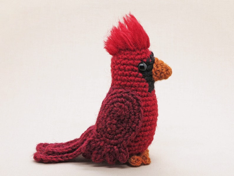 May include: A crocheted cardinal bird toy with a red body, black eyes, and an orange beak. The bird is sitting on a white surface.